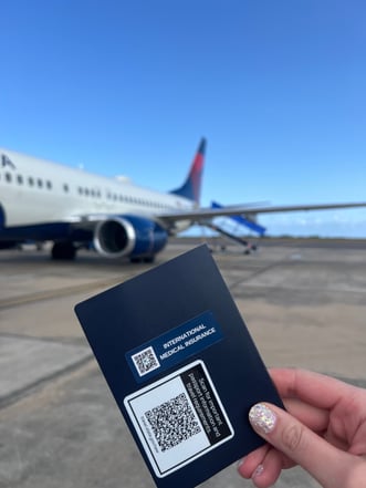 A hand holds up a passport with a MissionSafe Traveler's Toolbox sticker on the back. The person is on a tarmac. A plane is in the background.