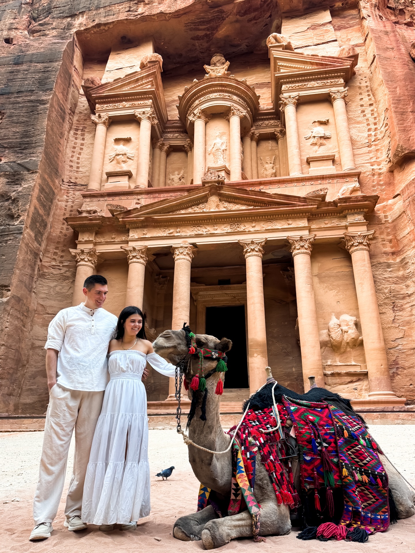 Travel Influencer Adventuring Cam and her husband stand next to a camel in front of the Treasury in Petra, Jordan.