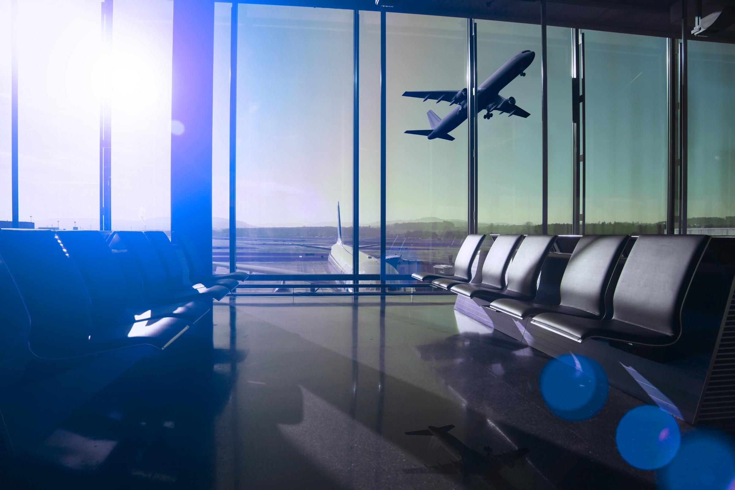 looking out a window at the airport. Terminal seats are in the foreground, the tarmac is in the background. Sun coming through window. Blue overlay.