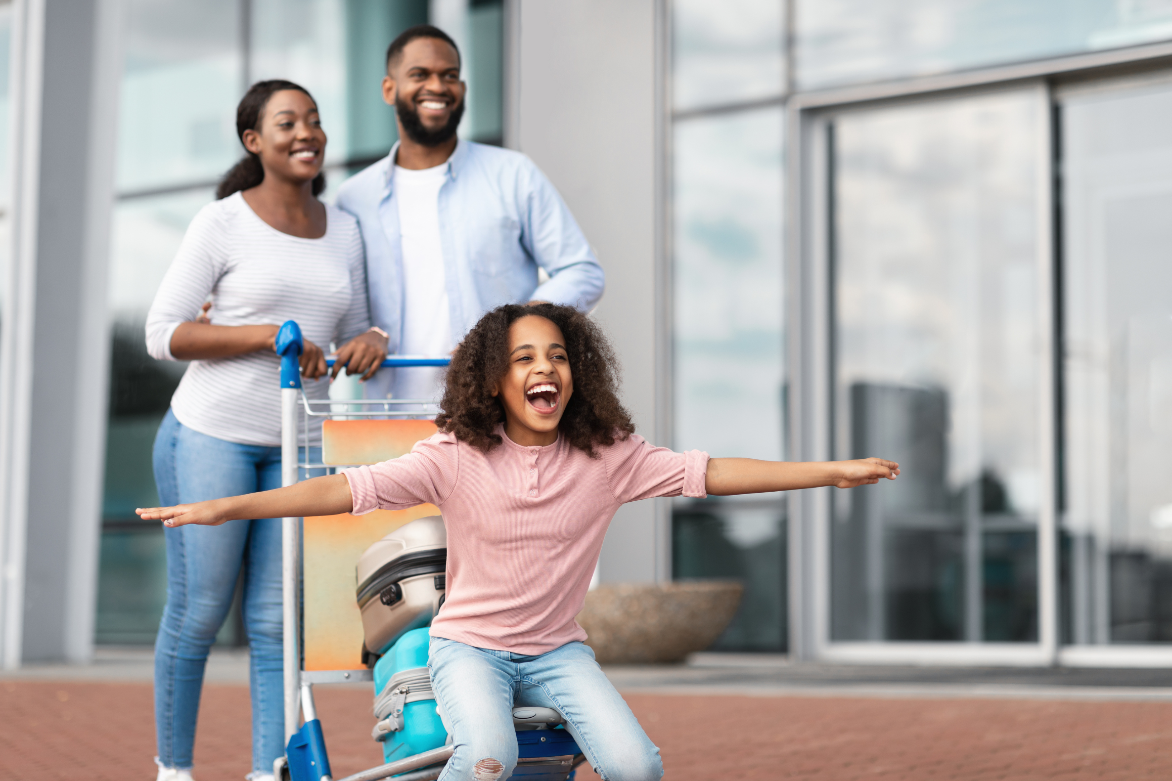 A joyful family outside an airport. A young girl sits on a luggage cart with her arms wide open, expressing excitement and happiness.